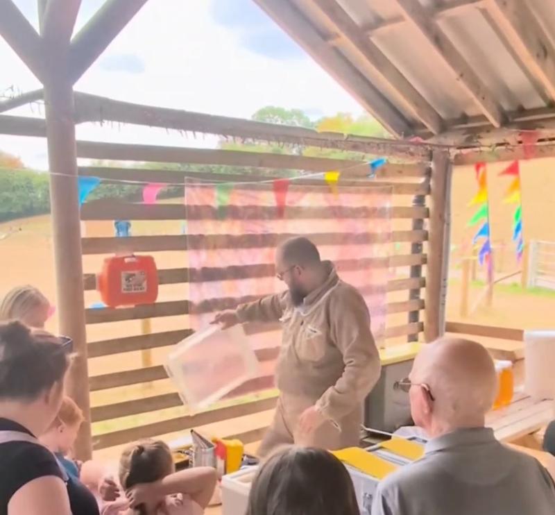 Beekeeper Markus Cox holding up a frame of honeycomb while talking to a seated group in an outdoor teaching shelter